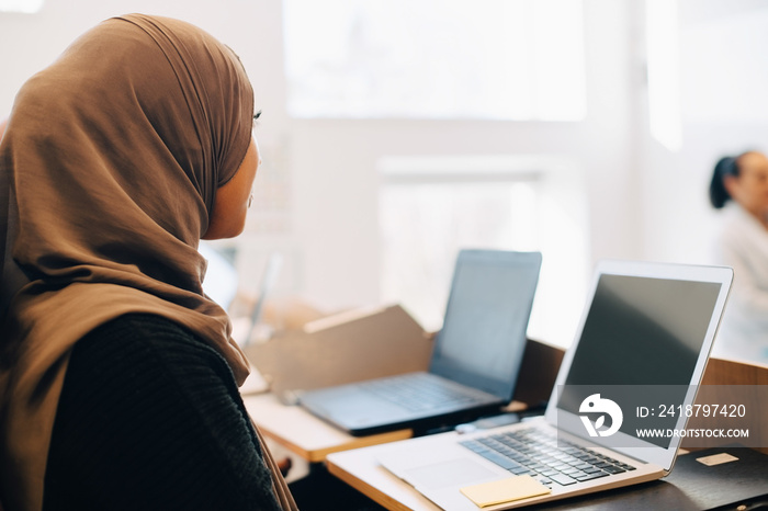 Side view of female student wearing hijab using laptop at desk in classroom