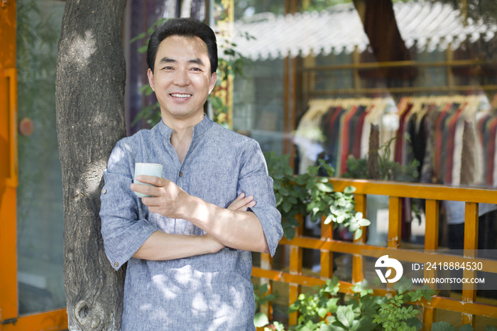 男性店主拿着茶杯站在店外