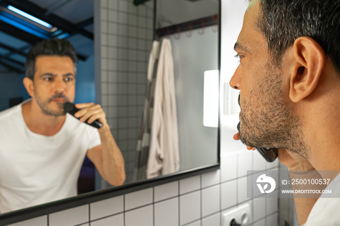 handsome man is shaving his beard with trimmer machine in front of bathroom mirror