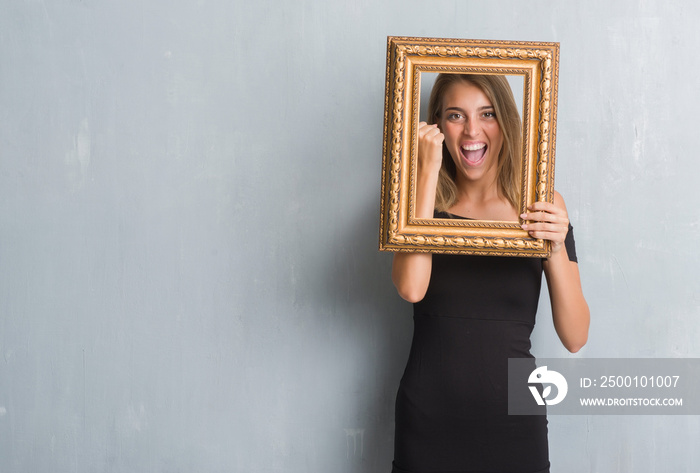 Beautiful young woman over grunge grey wall holding vintage frame screaming proud and celebrating victory and success very excited, cheering emotion