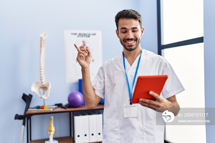 Young hispanic physiotherapist man doing online appointment smiling happy pointing with hand and finger to the side