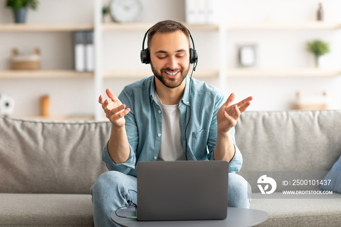 Happy young guy in headset making video call on laptop at home, talking to camera