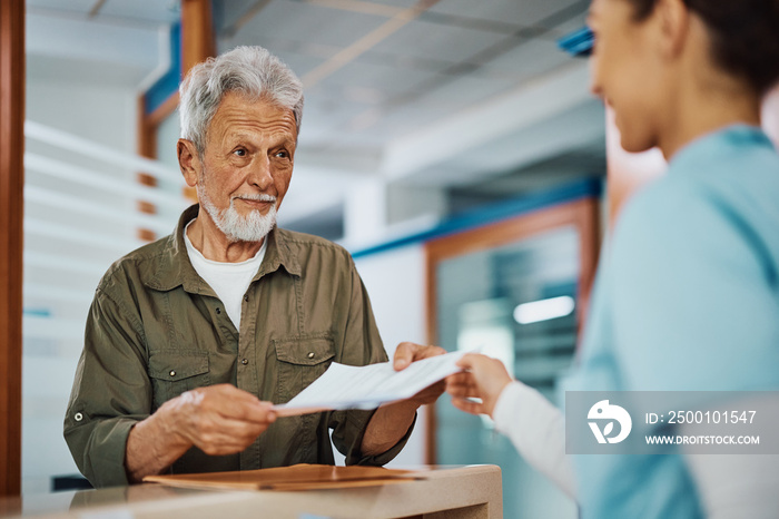 Elderly man receiving his medical documents from nurse at reception desk at doctor’s office.