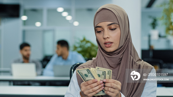 Serious concentrated eastern muslim girl in hijab office worker manager or accountant holding bundle of dollars in hands carefully counting cash paper money on payday keeps records of company income