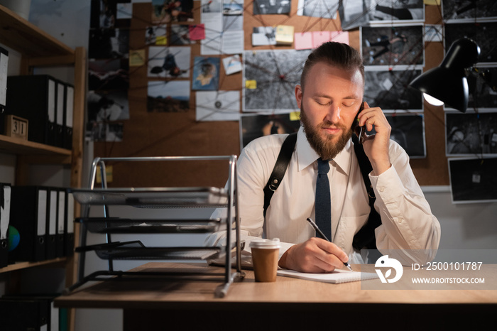 A professional detective works on a murder in the office at night. Sitting at the table takes a call from a witness and writes down the testimony in a notebook.