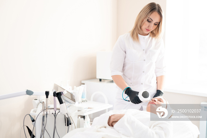 Young cosmetologist in surgical gloves providing laser treatment to mature woman on procedure table in beauty salon