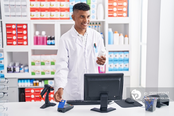 Young latin man pharmacist using credit card and data phone holding pills bottle at pharmacy