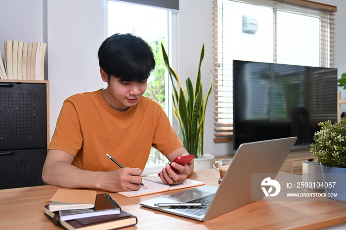 Happy asian man holding smart phone and making notes on notebook.