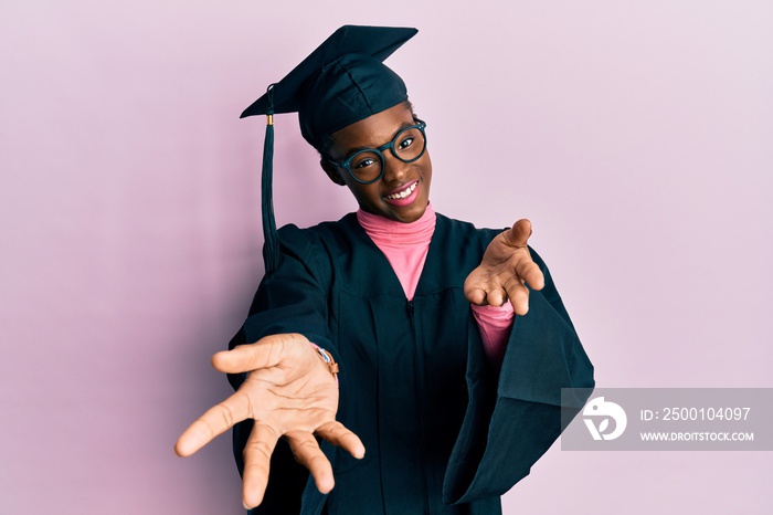 Young african american girl wearing graduation cap and ceremony robe smiling cheerful offering hands giving assistance and acceptance.