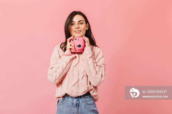 Smiling dark-haired lady in stylish sweater poses with pink camera on isolated background