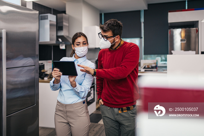 Young man talking with saleswoman about refrigerator he wants to buy. They are both with face protective masks. Pandemia, Covid-19 concept.