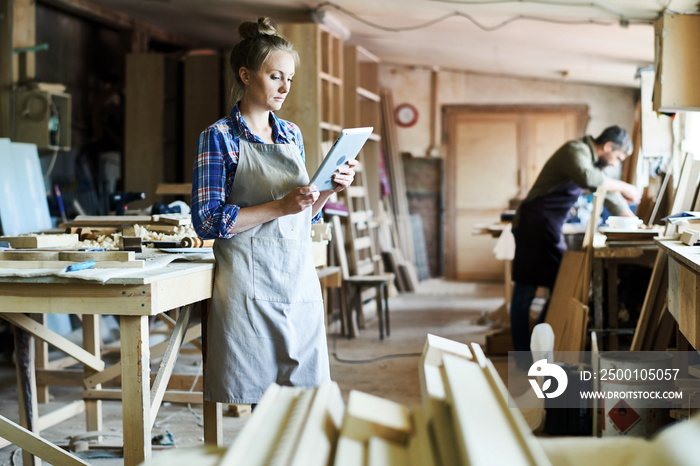 Full length portrait of concentrated young craftswoman in apron standing at spacious workshop and using digital tablet