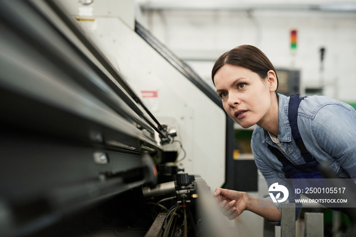 Content skilled brunette woman in workwear watching lathe machine processing and adjusting it at factory