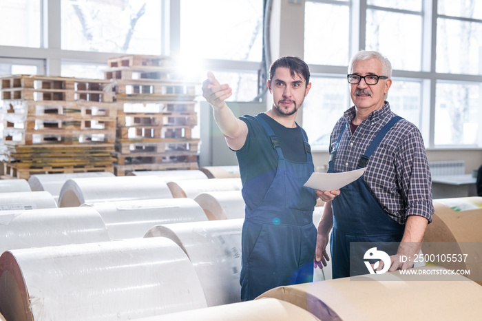 Senior and young warehouse workers of printing plant standing among paper rolls and discussing delivery