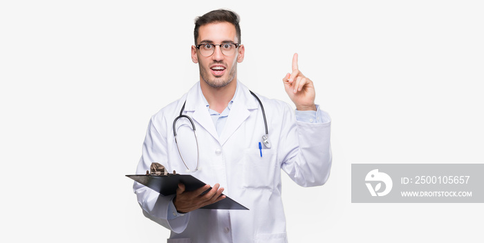 Handsome young doctor man holding a clipboard surprised with an idea or question pointing finger with happy face, number one