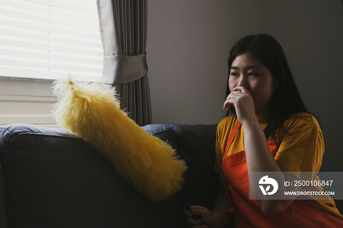 Asian woman doing housework uses dusting sticks to dust the window blinds that are so dirty and dusty that she has to cover her nose with toilet paper to prevent allergies and sneezes.