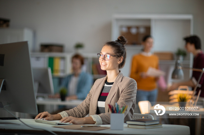 Happy young office manager or designer sitting by desk in front of computer screen while working with online content