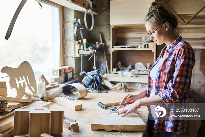 Profile view of confident young craftswoman in eyeglasses measuring wooden plank with pencil and ruler while standing at table in messy workshop