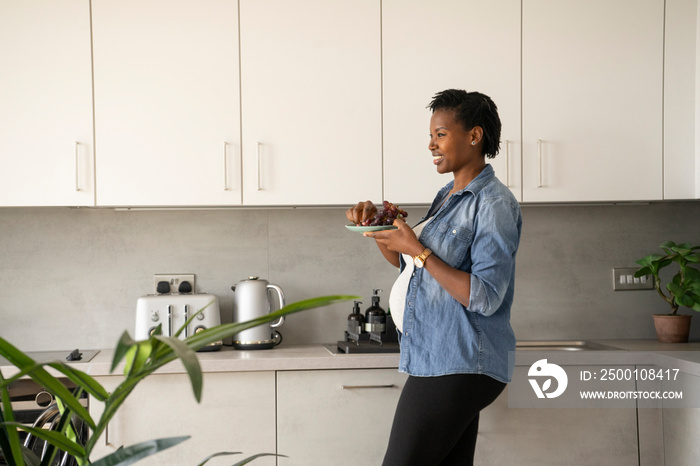 Smiling pregnant woman holding plate with red grapes in kitchen