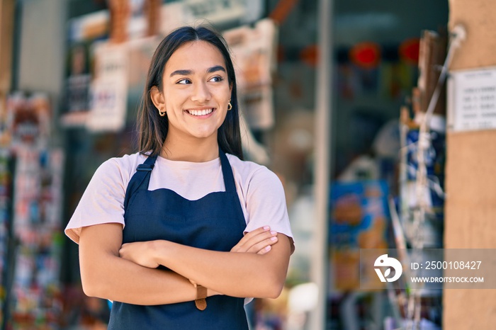 Young latin shopkeeper girl with arms crossed smiling happy at the press shop