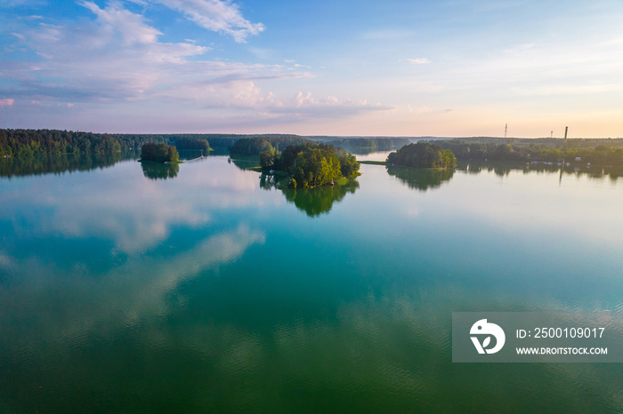 Aerial view of the lake surrounded by the green forest. Blue sky, mirror image of white clouds in the water.