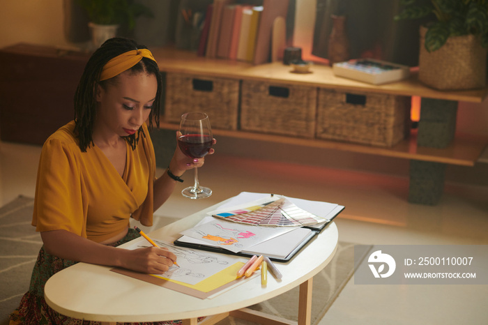 Creative Black woman drinking glass of red wine when drawing fashion sketches for new season