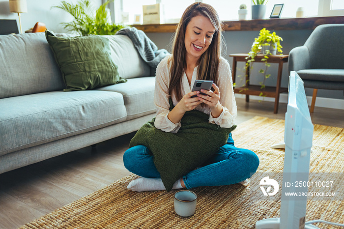 Woman dressed in beige sweater regulating heating temperature with a modern wireless thermostat and smart phone at home. Synchronization of thermostat with mobile devices concept