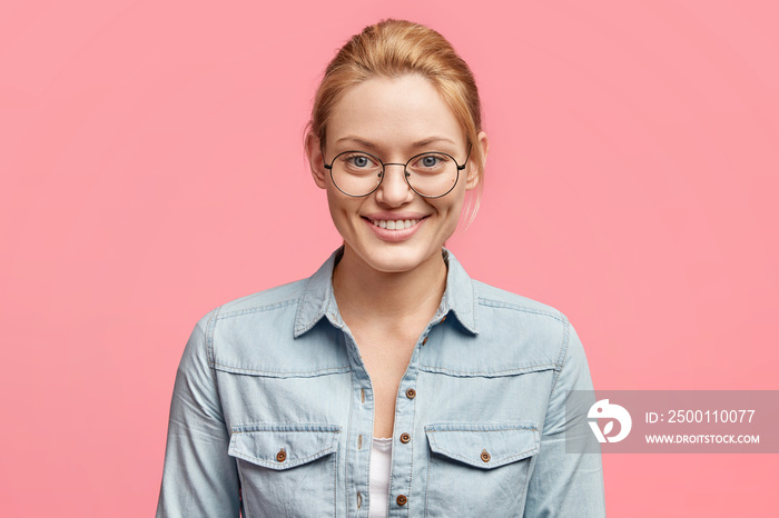 Portrait of pleased attractive smiling female dressed in fashionable denim jacket, sees something appealing in shop, isolated over pink studio background. Cheerful blonde young European woman