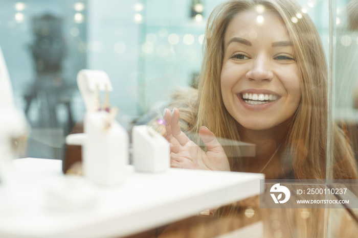 Selective focus of happy young girl looking through glass at new accessories in jewelry store. Lady laughing and choosing new jewelry before purchasing. Concept of shopping and happiness.