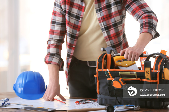 Male builder with tools in bag and house plan in office