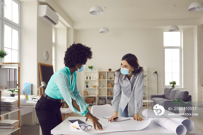 Coronavirus pandemic, safety, return to workplace after end of lockdown and quarantine. Two young creative designers and business women in face masks meeting in office studio and working on draft plan