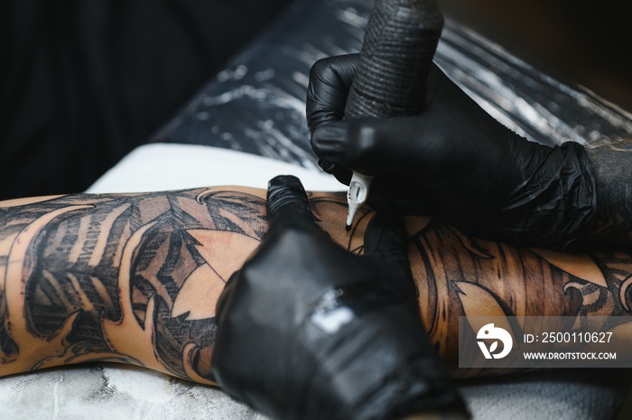 Cropped close up of a bearded tattoo artist working at his studio tattooing sleeve on the arm of his male client. Man getting tattooed by professional tattooist