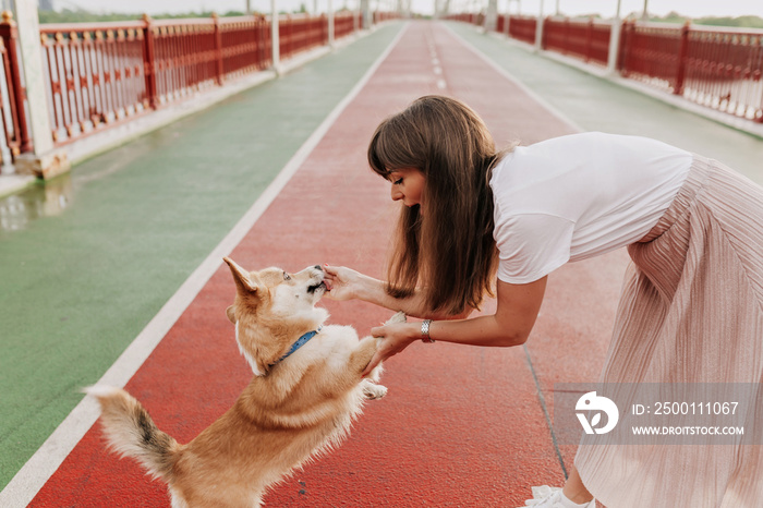 View of cheerful woman on the street with her dog. Woman in white t-shirt and pink skirt playing with corgi dog