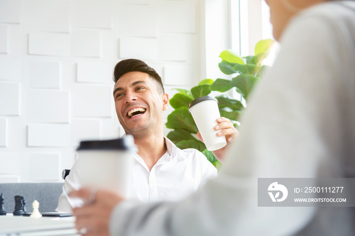 A man in casual white shirt is laughing and drinking coffee while having a meeting with his colleagues in the greenery white office pantry.