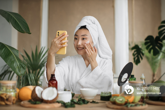 Happy asian woman holding bowl applying white mask homemade moisturizing and nourishing face mask or cream making mixture with natural cosmetic products.