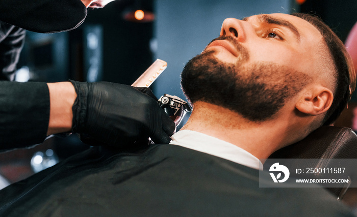 Young man with stylish hairstyle sitting and getting his beard shaved in barber shop