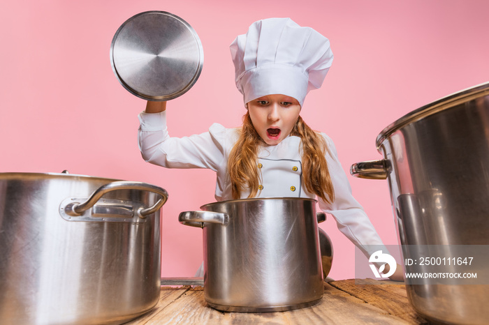 Close-up cute little girl in white cook uniform and huge chef’s hat at kids kitchen with big pans isolated on pink studio background.
