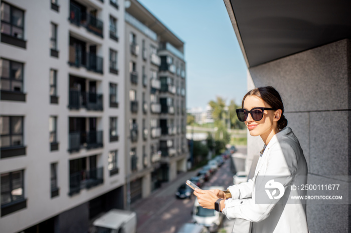 Young business woman in white suit and sunglasses on the balcony of the apartment in the modern residential area