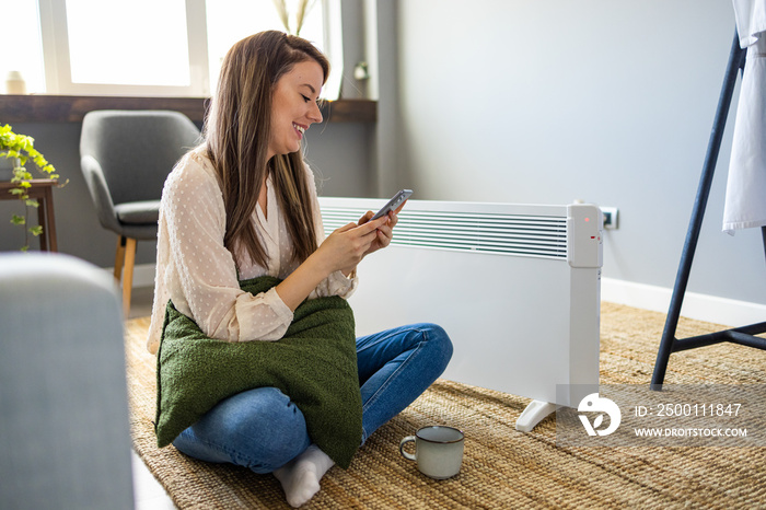 Woman regulating heater temperature. woman in beige blouse in the modern house in sunny winter day sitting near couch and white electric oil radiator using smartphone app for temperature control.