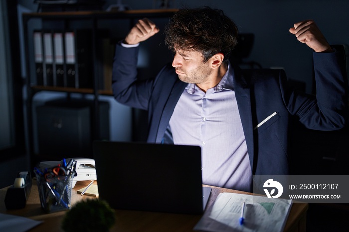 Hispanic young man working at the office at night showing arms muscles smiling proud. fitness concept.