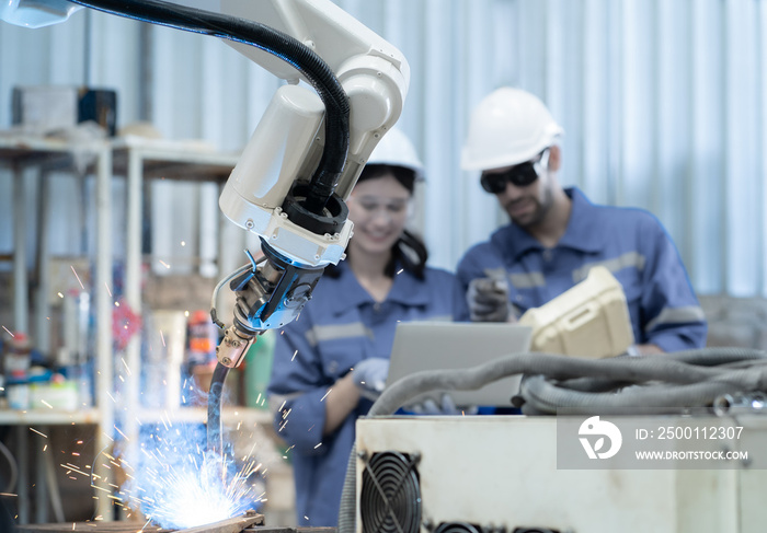 Industrial engineer using laptop computer to control robotic welder arm operation in modern automation factory. Technician monitoring robot machine arm does automated steel welding full of fire sparks