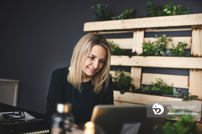 pretty blond young woman is sitting in an ecological office with lots of plants and is working on her laptop and is wearing a green sweater, concept sustainability and environment today