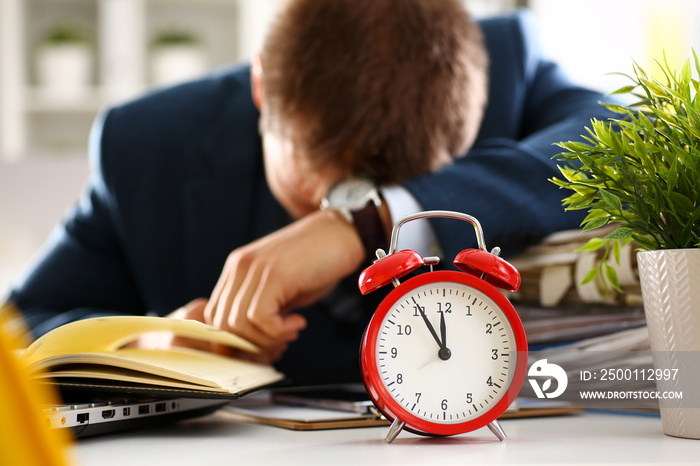 Red alarm clock shows late time closeup and tired office male clerk in suit take nap on table workplace full of exam papers. Career frustration freelance employment fail study problem low energy down