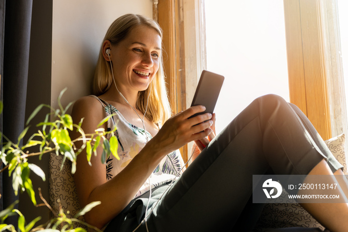 smiling young woman sitting on sunny window sill and using smartphone at home