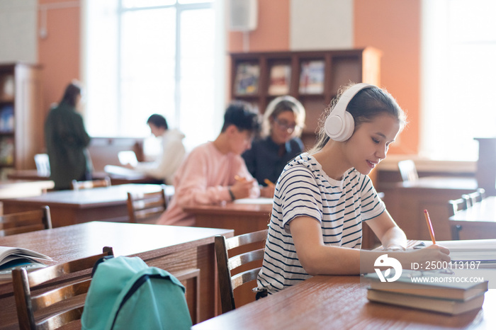 Contemporary college student with headphones writing down plan of seminar in notepad while sitting in library