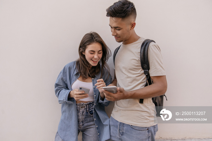 Young pair looking at phone against light wall background. Attractive brunette-haired boy in beige t-shirt showing something at phone to girlfriend, smiling and wearing denim clothes