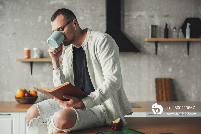 Portrait of a man drinking coffee wearing eyeglasses and reading a book while resting in kitchen. Coffee break.