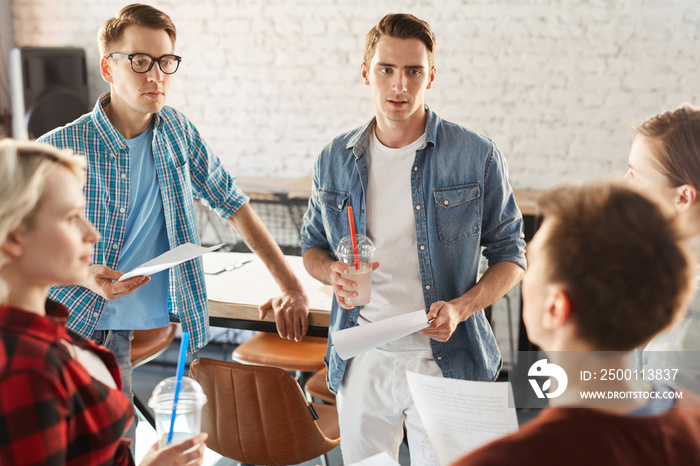 Group of creative young professionals discussing ideas while collaborating during meeting in modern office or cafe, focus on handsome young man holding smoothie