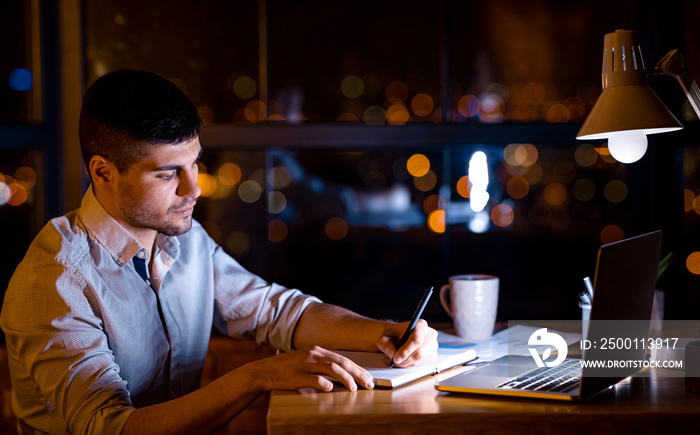 Freelancer Guy Taking Notes Sitting At Laptop Working Remotely Overnight In Home Office