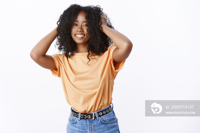 Attractive carefree young female african-american wearing trendy orange t-shirt dancing happily smiling touching afro curly haircut delighted, feeling fluffy hair, standing white background